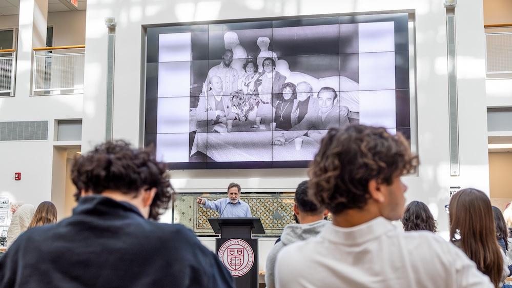 Prof. Roger Gilbert introduces the Ammons Reading Series to an audience of students and faculty in the Klarman Hall Groos Family Atrium, displaying a slideshow of historical photos from the era of Ammons attendance at undergraduate open mic events in the former Temple of Zeus Cafe, Goldwin Smith Hall.
