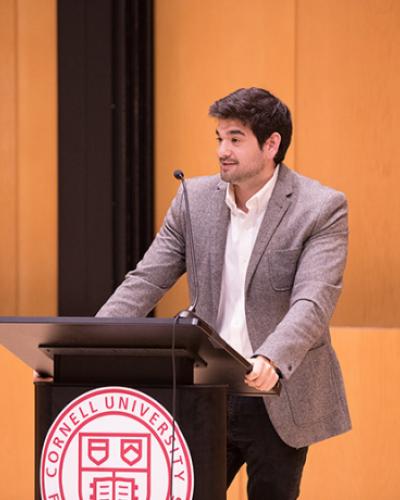 Christopher Berardino reading at podium decorated with Cornell seal
